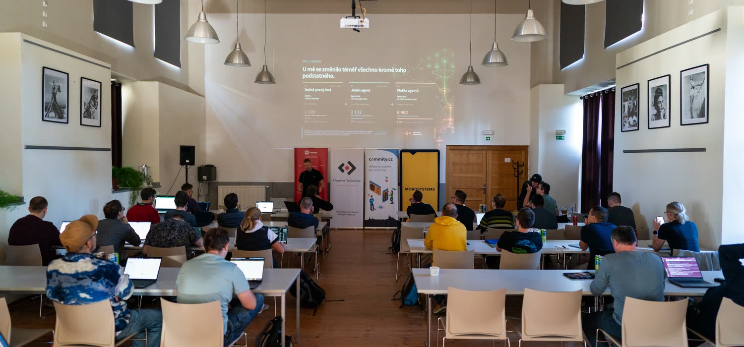 Pohled přes publikum na přednášejícího v konferenčním sále.