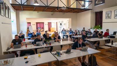 Účastníci sedí u stolů s notebooky v prosvětleném konferenčním sále.