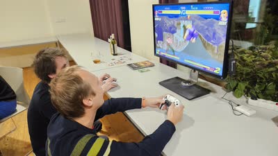 Dva účastníci s ovladači hrají proti sobě bojovou hru na monitoru.