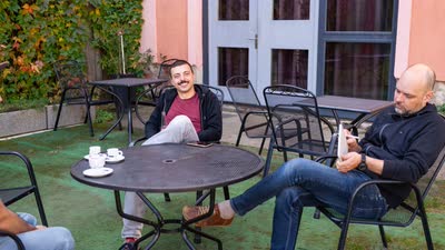 Dva účastníci odpočívají u stolku na terase; jeden se dívá do telefonu.